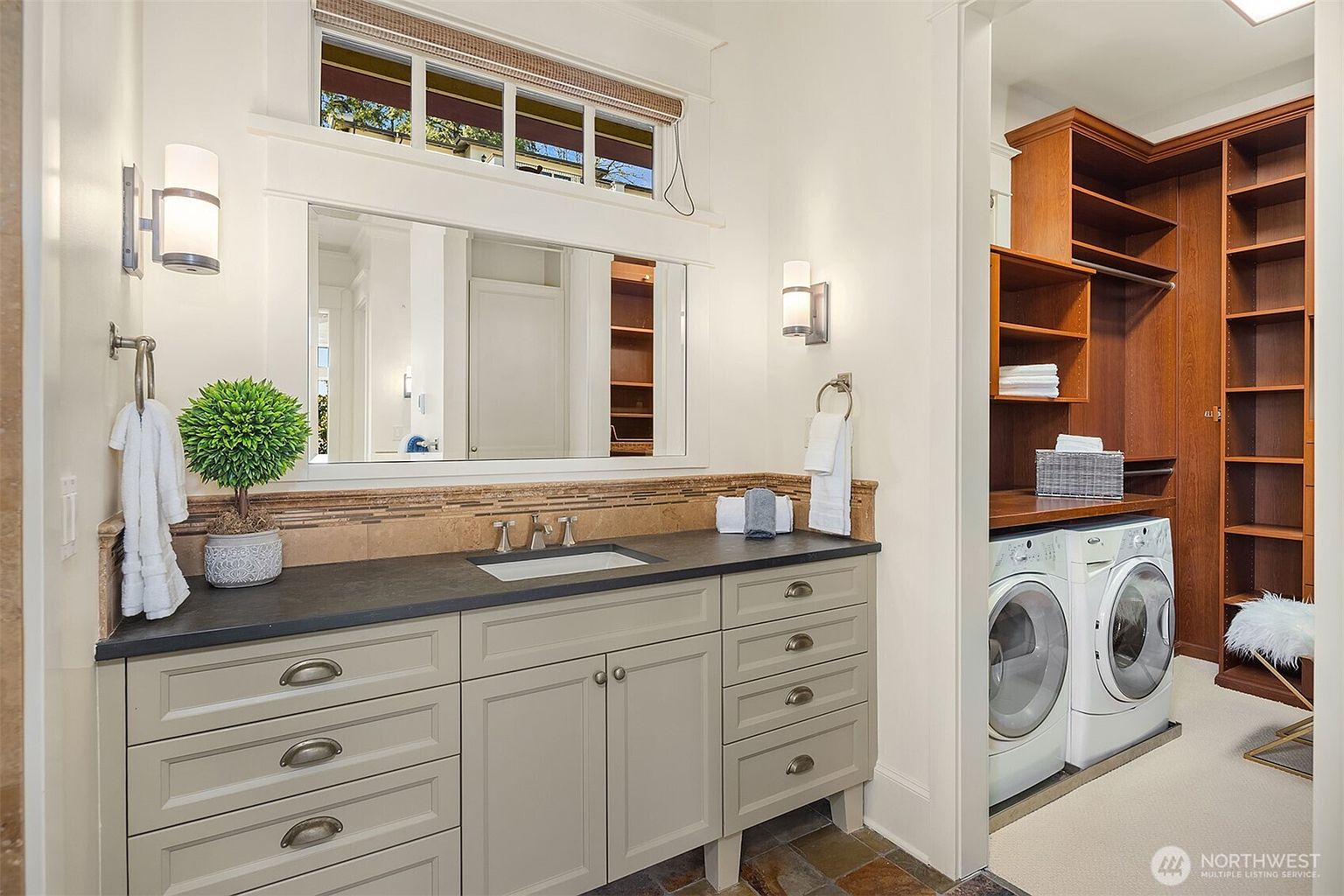 This elegant primary bathroom features a light gray vanity with a dark stone countertop, complemented by a large mirror and classic wall sconces. To the right, an open-concept laundry area is seamlessly integrated with custom wooden shelving and a front-loading washer and dryer set. The space is bright and functional, blending sophisticated design with practical utility.