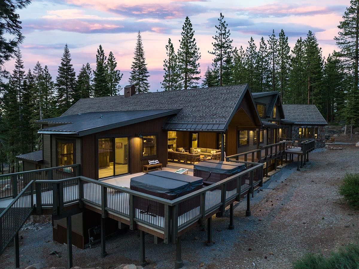 This image showcases the rear exterior of a luxurious mountain home at dusk, featuring a spacious deck with multiple hot tubs and outdoor seating. The architecture blends seamlessly with the surrounding pine forest, and the warm interior lighting creates an inviting ambiance. The property exudes a sense of privacy and relaxation.