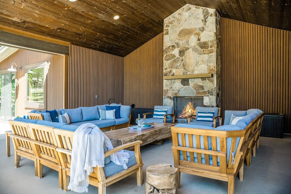 This image showcases a beautifully designed outdoor living space with a rustic yet modern aesthetic. The area features comfortable wooden furniture with blue cushions arranged around a unique wooden coffee table and a stone fireplace, creating a cozy and inviting atmosphere. The space is enclosed with wooden slat walls and a wooden ceiling, enhancing the natural and warm feel of the area.