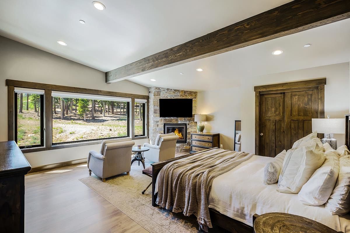 This is a primary bedroom featuring a king-size bed with white linens and a throw blanket. The room has a stone fireplace with a mounted television above it, two armchairs, and a large window offering a view of the trees outside. The style is rustic and luxurious, with wooden beams and trim adding warmth and character.