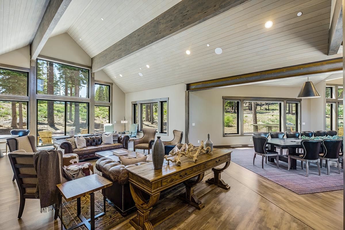 This is an interior shot of a spacious living room and dining area featuring hardwood floors, exposed wooden beams, and large windows that offer natural light and views of the surrounding trees. The living area includes leather sofas and armchairs, while the dining area has a large table with black chairs, creating an inviting and luxurious atmosphere. The room is decorated with a mix of modern and rustic elements, enhancing its appeal.