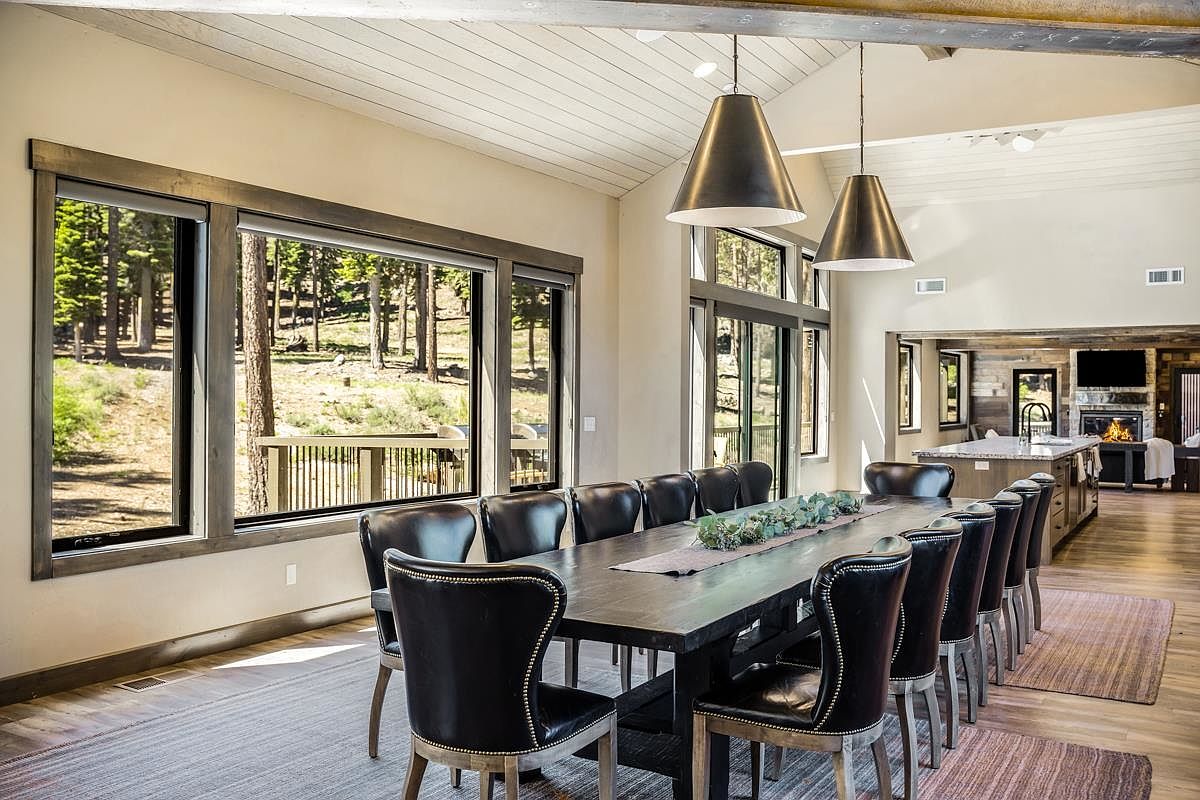 This is an interior shot of a dining room featuring a long, dark wood dining table with black leather chairs. Large windows offer a view of a wooded area, and modern pendant lights hang above the table. The room has a neutral color palette with wood accents, creating a sophisticated and inviting atmosphere.