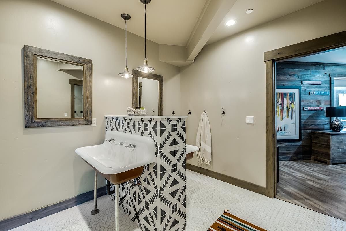 This is a stylish guest bathroom featuring a unique double sink vanity with a geometric black and white design. The sinks are vintage-style with exposed plumbing, complemented by rustic wood-framed mirrors and pendant lighting. The white hexagonal tile flooring and neutral wall color create a clean and inviting space.