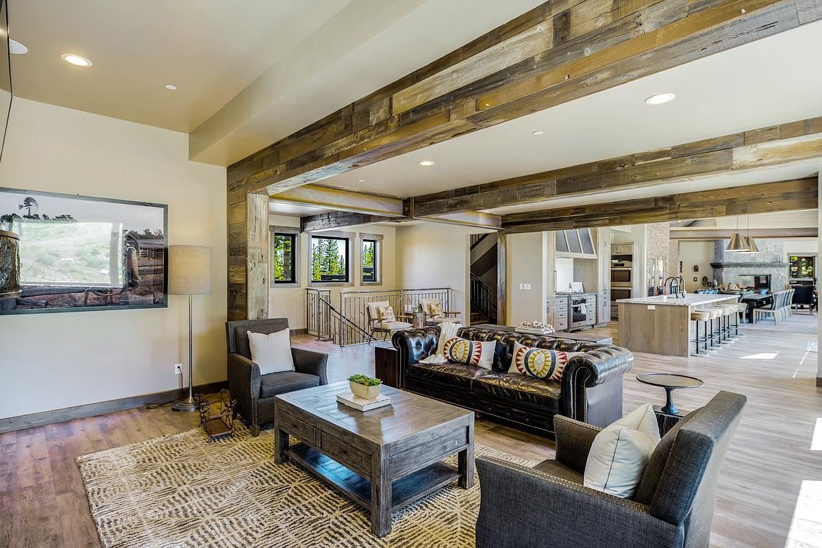 This is an interior shot of a living room featuring rustic wooden beams, hardwood floors, and a neutral color palette. The room is furnished with a leather sofa, two armchairs, and a wooden coffee table, creating a cozy and inviting atmosphere. The open floor plan seamlessly connects the living room to the kitchen and dining area, enhancing the sense of spaciousness.