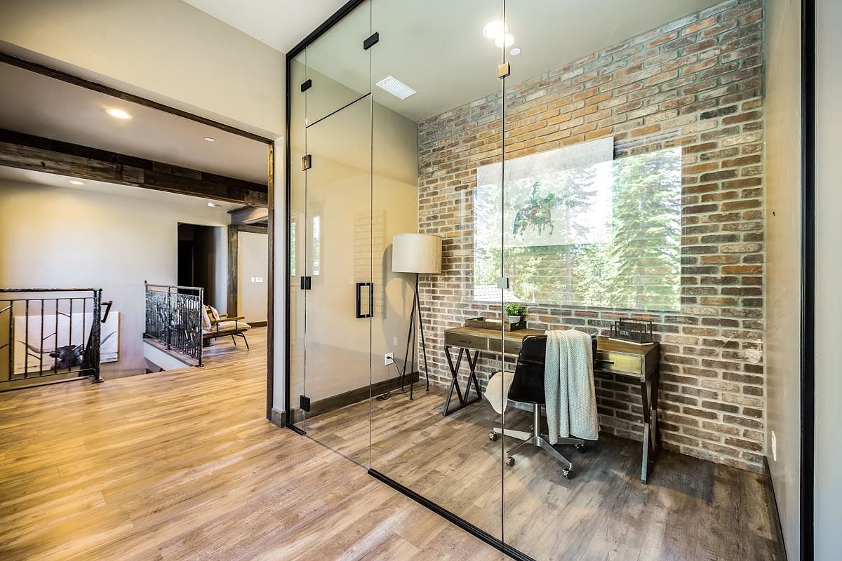 This is an interior shot of a modern office space featuring a glass-enclosed room with a brick accent wall. The office includes a desk, chair, and a floor lamp, creating a functional and stylish workspace. The flooring appears to be wood laminate, and the overall design is contemporary with an industrial touch.
