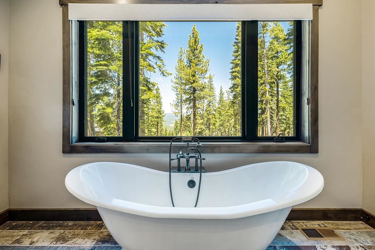 This image showcases a luxurious primary bathroom featuring a freestanding white bathtub positioned in front of a large window. The window offers a serene view of tall evergreen trees under a clear blue sky, creating a spa-like atmosphere. The bathroom's design incorporates natural wood trim and neutral wall colors, enhancing the sense of tranquility and connection with nature.