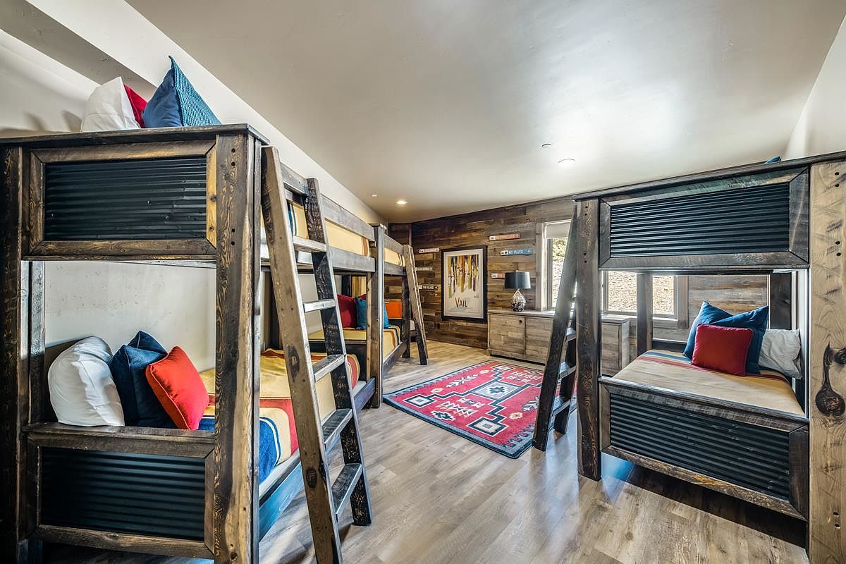 This is a bedroom featuring multiple bunk beds, constructed with a rustic wood and corrugated metal design. The room has a cozy, cabin-like feel, enhanced by the wood-paneled wall and a colorful area rug. The beds are adorned with pillows and blankets, creating an inviting space for guests.