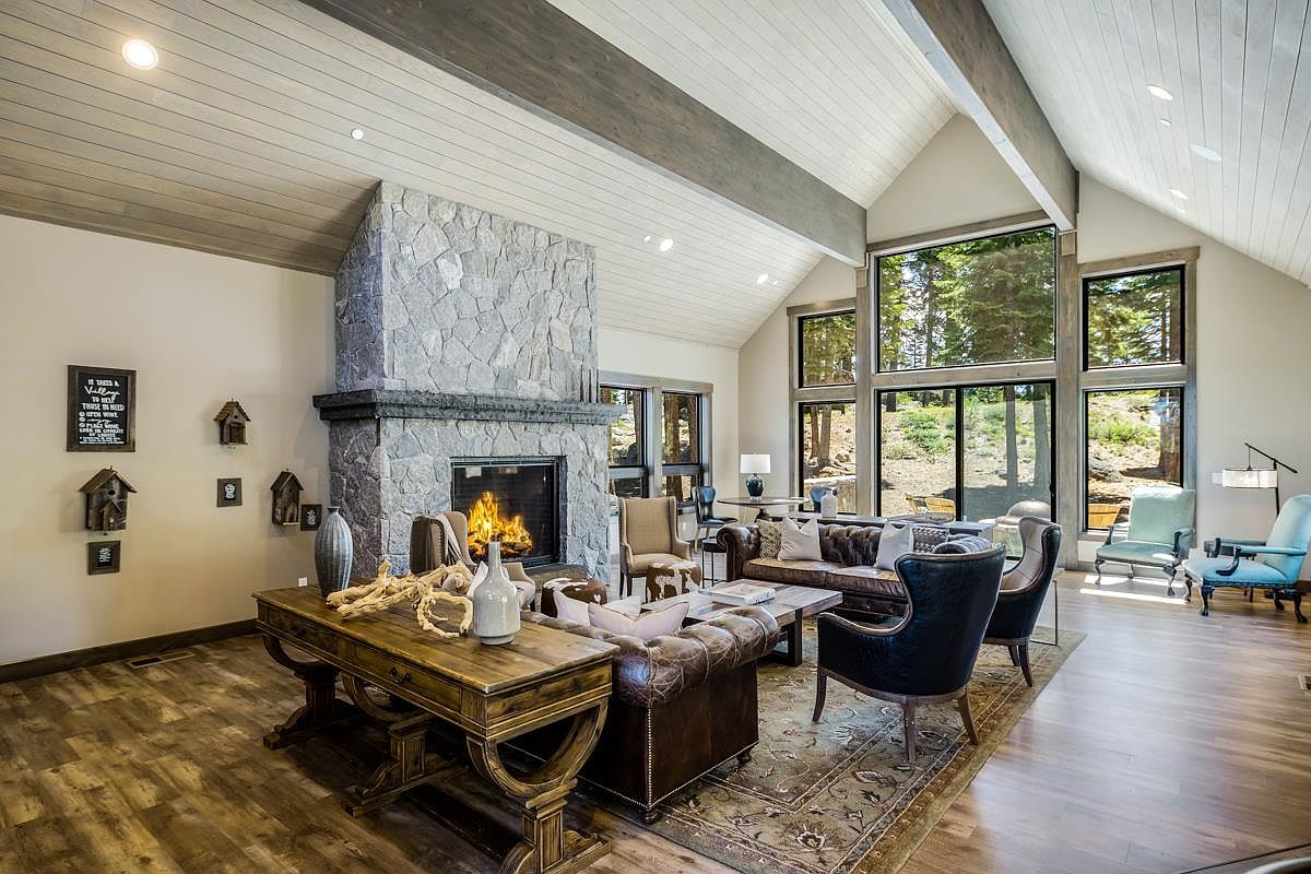 This is an interior shot of a living room featuring a stone fireplace, wooden floors, and large windows that offer a view of the outdoors. The room is furnished with leather sofas, armchairs, and a wooden desk, creating a cozy and inviting atmosphere. The high ceilings with exposed beams add to the room's spaciousness and character.