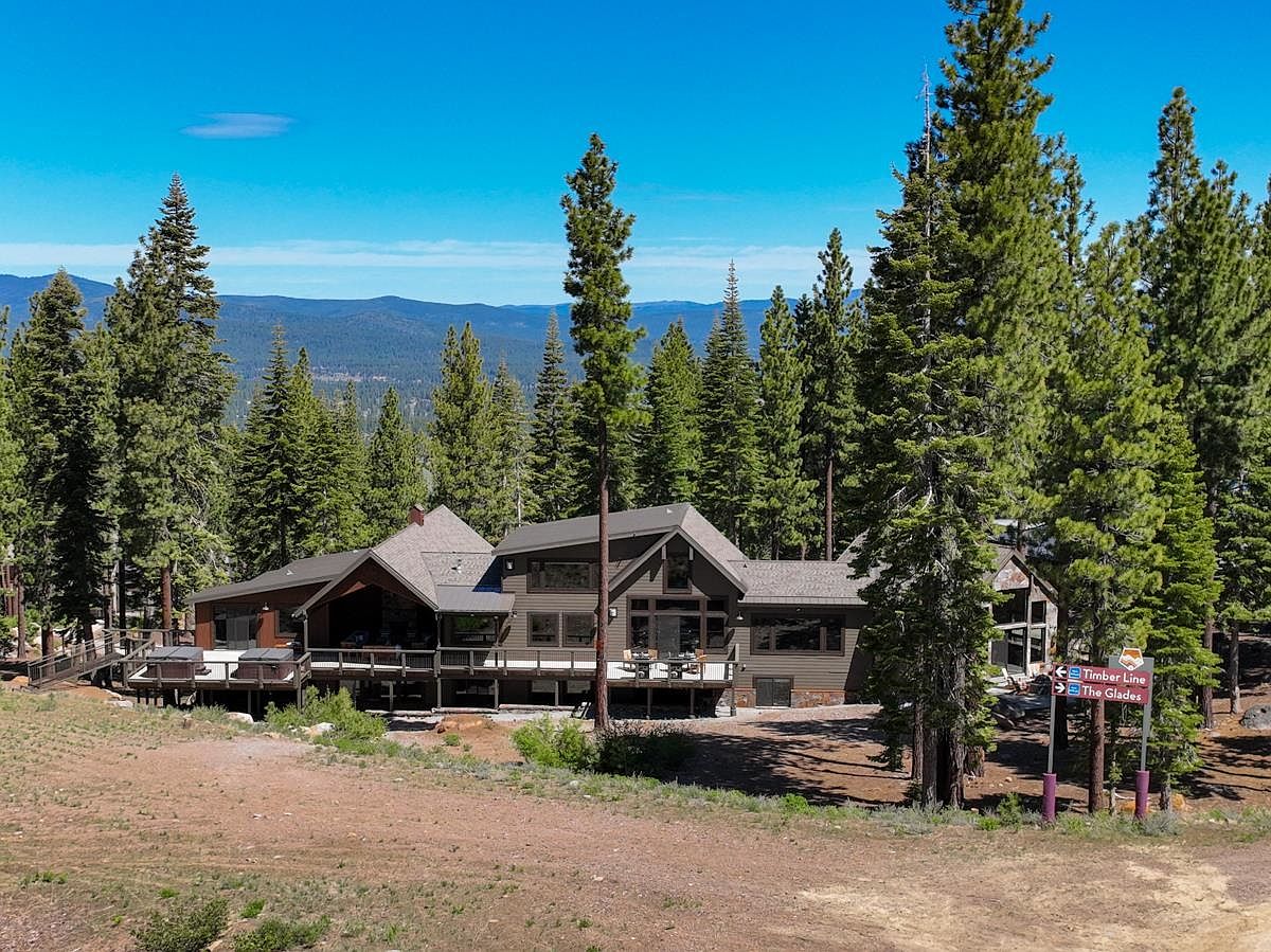 This aerial view showcases a large, modern mountain home nestled among tall evergreen trees. The house features a multi-level deck, a mix of wood and stone exterior, and a complex roofline. The surrounding landscape includes a dirt yard and distant mountain views, creating a sense of seclusion and natural beauty.