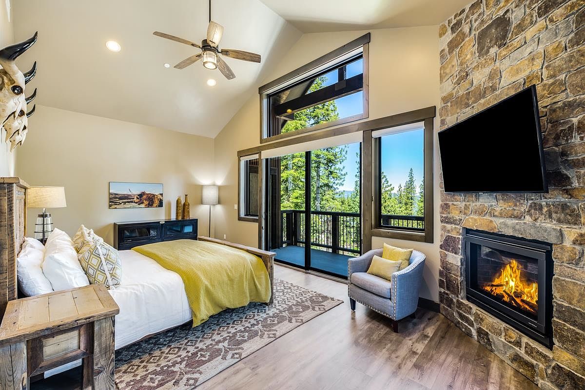 This is a primary bedroom featuring a stone fireplace with a mounted television, a king-size bed with a yellow throw, and a sitting area with a blue armchair. The room has a high vaulted ceiling with a ceiling fan and large windows that offer a view of the surrounding trees. The style is rustic and luxurious, creating a cozy and inviting atmosphere.
