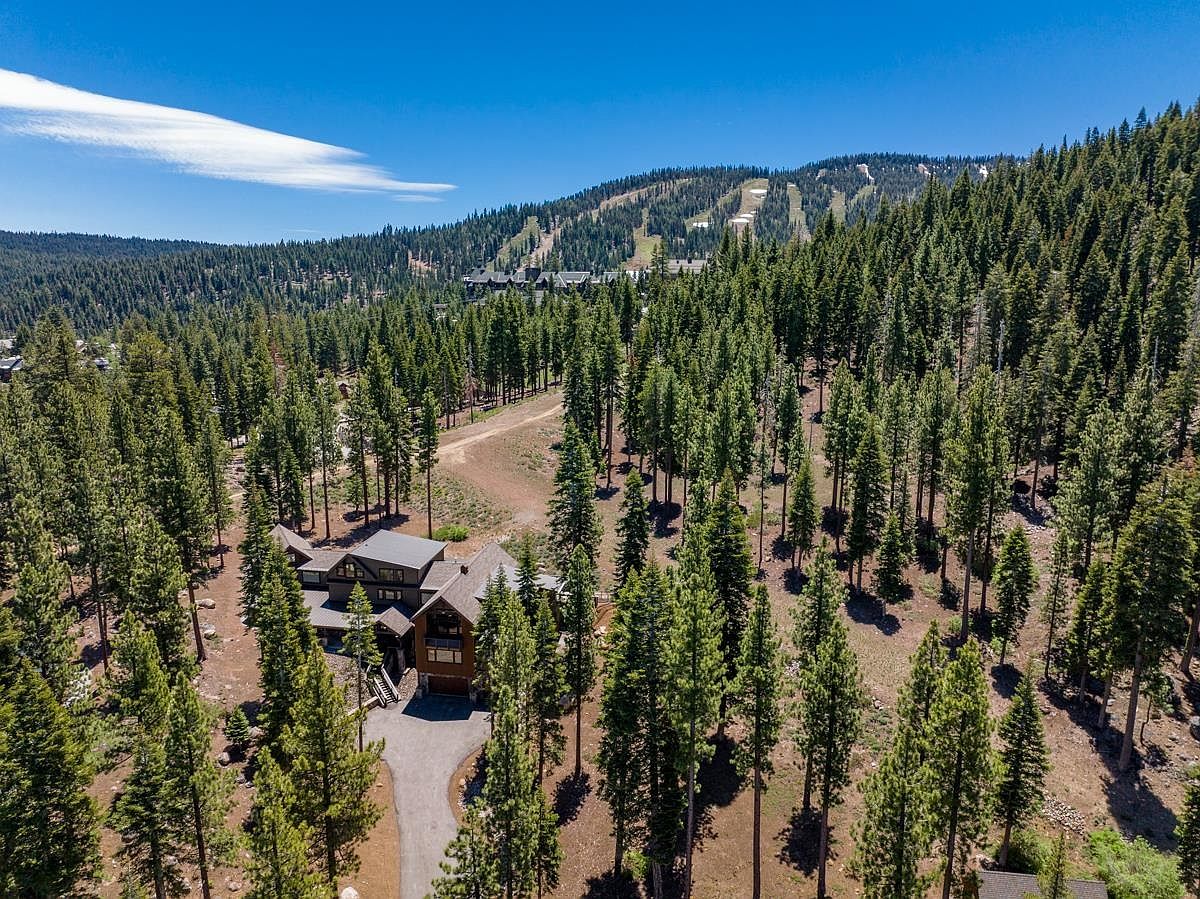 This aerial shot showcases a luxurious mountain home nestled among tall evergreen trees. The house features a multi-level design with a gray roof and a paved driveway leading to the entrance. In the background, a ski resort is visible on the mountainside, suggesting a prime location for outdoor enthusiasts.