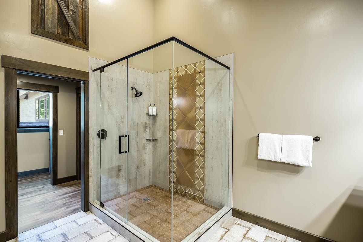 This is a well-lit primary bathroom featuring a modern glass-enclosed shower with decorative tile accents. The shower has a black shower head and handle, and a built-in shelf for toiletries. The bathroom floor is tiled, and a towel rack with white towels is mounted on the wall, creating a clean and inviting space.