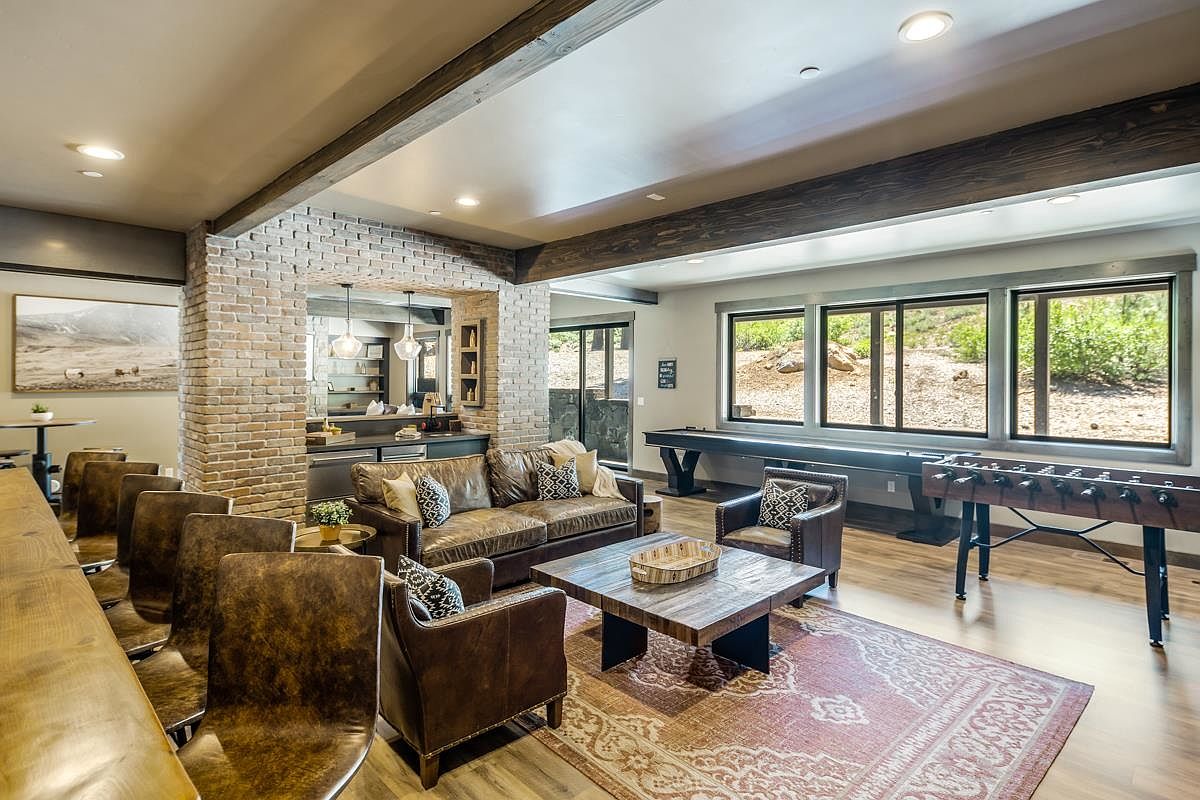 This is an inviting living room featuring a comfortable leather sofa and armchairs arranged around a rustic wooden coffee table on a patterned rug. The room includes a bar area with brick accents and a view of the outdoors through large windows, which also illuminate a shuffleboard table and foosball table. The space is designed for relaxation and entertainment.