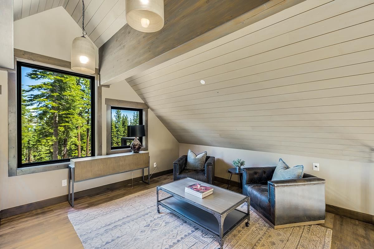 This interior shot showcases a cozy living room area with a sloped ceiling and wood paneling. Two armchairs flank a rustic coffee table, set upon a patterned rug. Large windows offer views of the surrounding trees, enhancing the room's natural light and serene atmosphere.