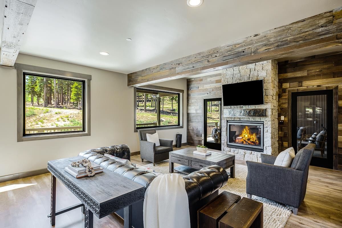 This is a cozy living room featuring a stone fireplace with a mounted television, comfortable seating, and rustic wood accents on the ceiling and walls. Large windows provide natural light and views of the surrounding trees, creating a warm and inviting atmosphere. The room is furnished with a leather sofa, armchairs, and a wooden coffee table, all arranged on a patterned area rug.