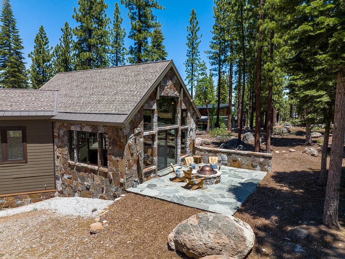 This image showcases the rear exterior of a house, highlighting a stone-clad facade with large windows and a gable roof. A stone patio with a fire pit and Adirondack chairs provides an inviting outdoor living space. The surrounding landscape features mature trees and natural ground cover, creating a serene and private setting.
