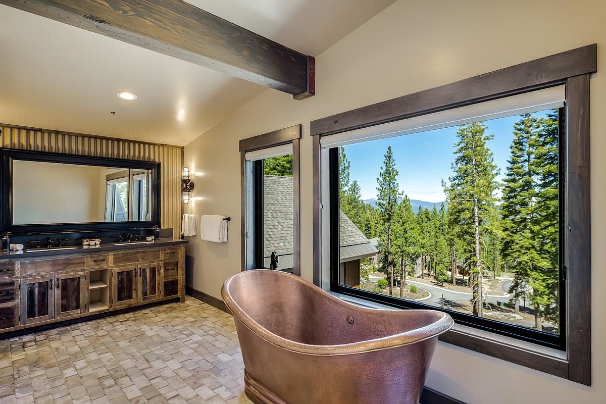 This is a primary bathroom featuring a copper freestanding bathtub positioned in front of a large window with a view of trees. The bathroom also includes a rustic wooden vanity with a large mirror and decorative lighting. The overall style is luxurious and rustic, creating a spa-like atmosphere.