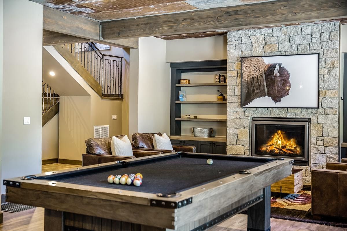This interior shot showcases a rustic living room with a stone fireplace and a large buffalo art piece above it. A pool table sits in the foreground, adding a recreational element to the space. The room features exposed wooden beams and a staircase in the background, contributing to a cozy and inviting atmosphere.