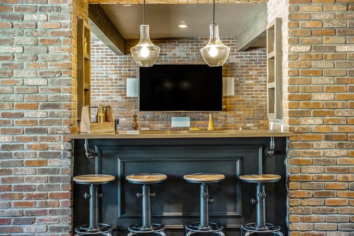 This interior shot showcases a stylish basement bar area, featuring a brick wall backdrop and pendant lighting. The bar itself is dark with a light countertop, complemented by four industrial-style stools. A television is mounted on the brick wall, creating a cozy entertainment space.