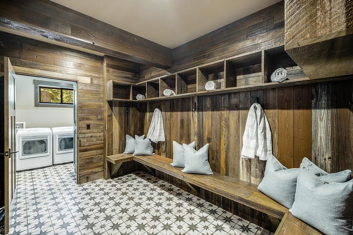 This is a well-organized laundry room featuring rustic wood paneling on the walls and ceiling, complemented by built-in shelving and a bench with soft pillows. The patterned tile floor adds a touch of elegance, while the modern washer and dryer are visible through an open doorway. The room exudes a cozy and functional atmosphere.