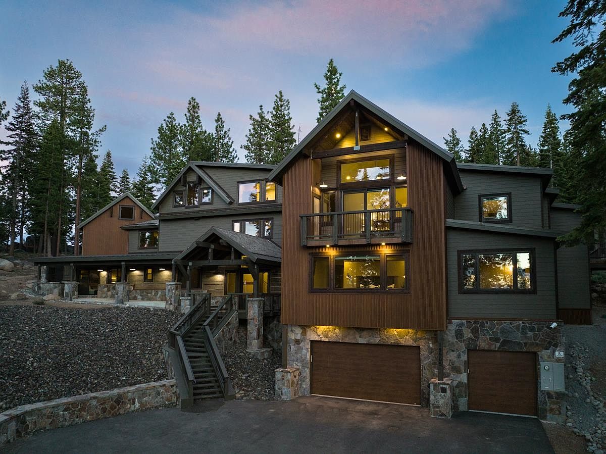 This is a front exterior view of a multi-level luxury home with a combination of wood and stone facade. The house features multiple windows, a balcony, and a prominent staircase leading to the main entrance. The property is surrounded by tall evergreen trees, creating a sense of privacy and seclusion.