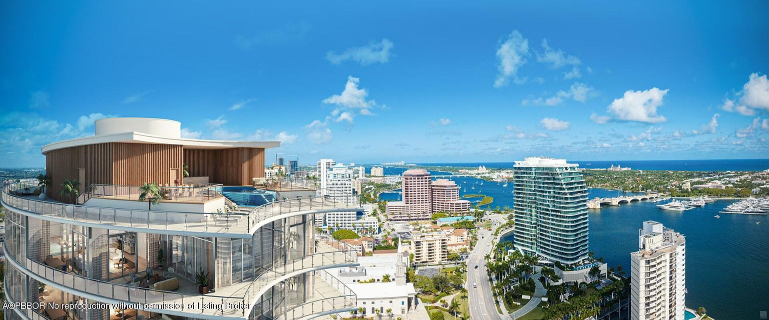 This aerial view showcases a luxurious high-rise condominium with expansive balconies and a rooftop pool, offering stunning panoramic views of the city skyline and waterfront. The building's modern architectural design is complemented by lush landscaping and nearby waterways filled with yachts, creating an impression of upscale coastal living. The clear blue sky and bright sunlight enhance the overall appeal of this prime real estate location.