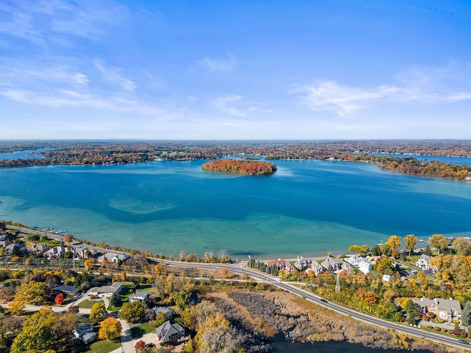 This aerial view showcases a stunning lakefront property with vibrant fall foliage surrounding the area. The crystal-clear blue lake features a small island covered in colorful trees, while luxury homes line the shore with well-maintained landscaping. A road winds along the waterfront, connecting the properties and offering scenic views.