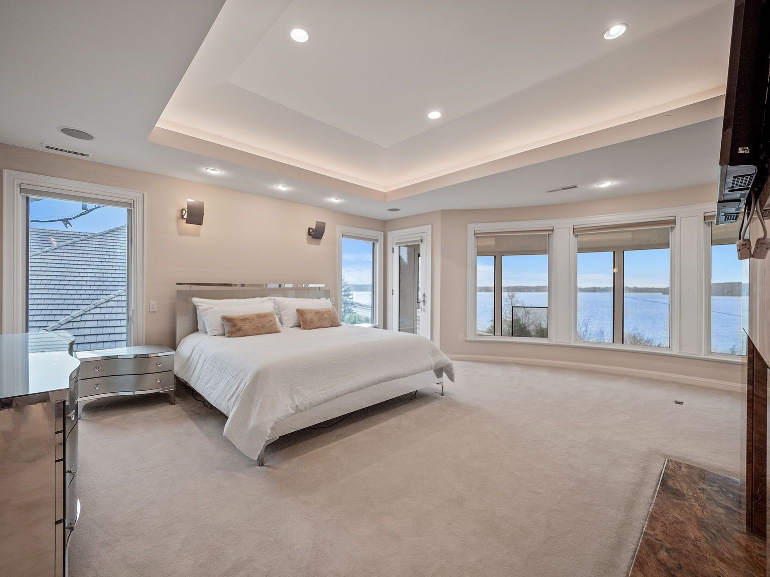 This is a spacious primary bedroom with a serene view of the lake. The room features a large bed with white linens, a mirrored nightstand, and a neutral color palette. Large windows provide ample natural light and showcase the waterfront scenery, enhancing the room's tranquil atmosphere.