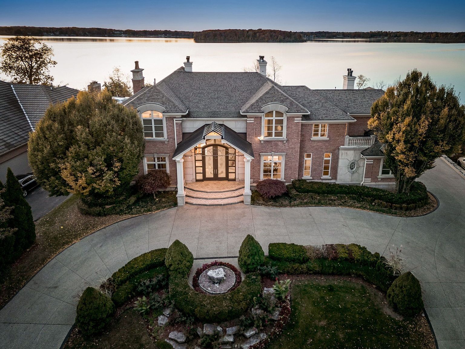 This aerial view showcases a grand brick mansion with a circular driveway and meticulously landscaped gardens, including a central fountain. The home features a prominent entryway with arched windows and a covered porch, complemented by a gray roof and multiple chimneys. The property overlooks a serene lake, enhancing its appeal as a luxurious waterfront estate.