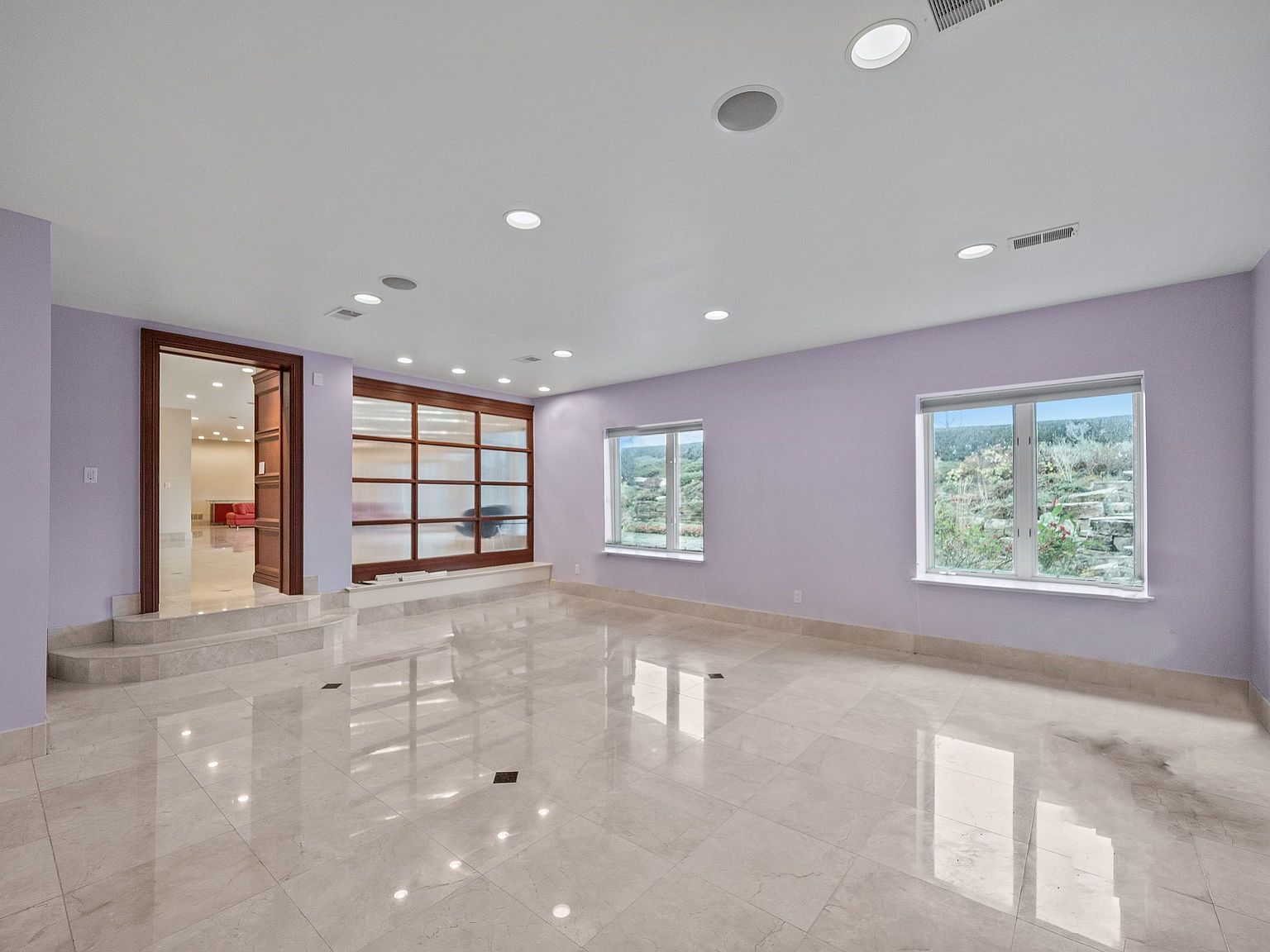 This is a spacious basement area featuring light purple walls, a glossy tiled floor, and recessed lighting. The room has two windows providing natural light and a large wooden-framed opening that leads to another room. The overall impression is clean and modern, with a touch of elegance.