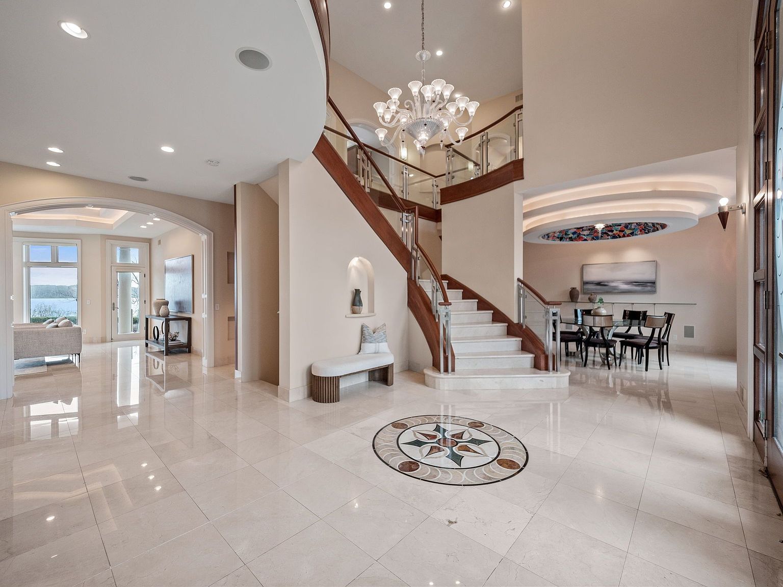 This is a grand foyer featuring a curved staircase with wood and glass railings, leading to an upper level. The flooring is light-colored tile with a decorative medallion at the center. A chandelier hangs above the staircase, and there's a dining area visible to the right, creating an elegant and welcoming entrance.