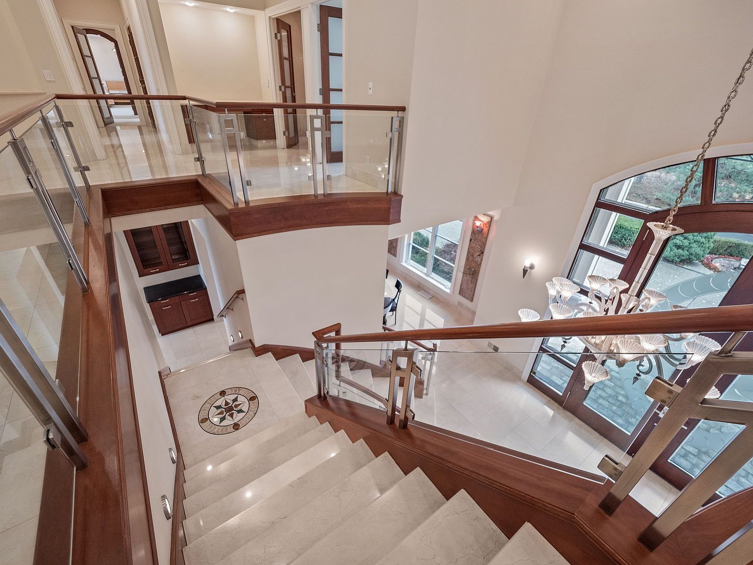 This interior shot showcases a grand staircase with marble steps, wood and glass railings, and a decorative medallion on the landing. The open design allows for ample natural light, highlighting the elegant chandelier and the view through the large windows. The overall impression is one of luxury and spaciousness.