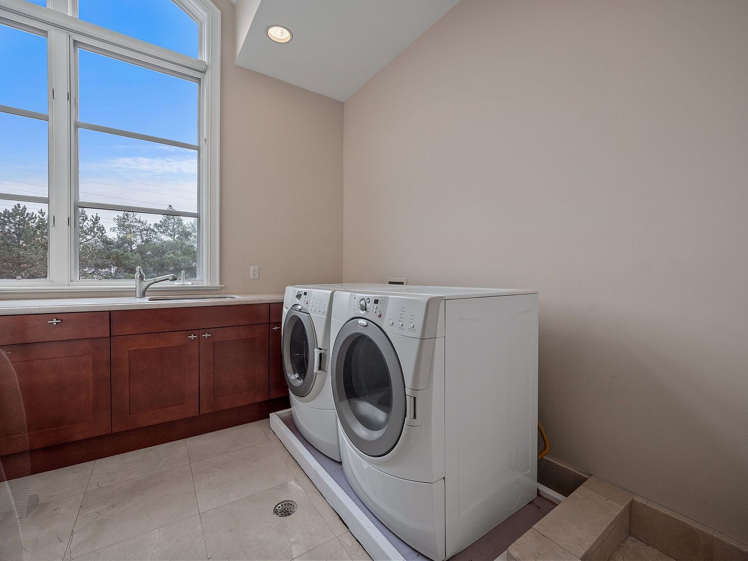 This is an interior shot of a laundry room featuring a pair of white front-loading washing machines and dryers. The room has dark wood cabinets with a light countertop and a sink under a large window that provides natural light. The flooring is light-colored tile, and the walls are painted in a neutral tone, creating a clean and functional space.