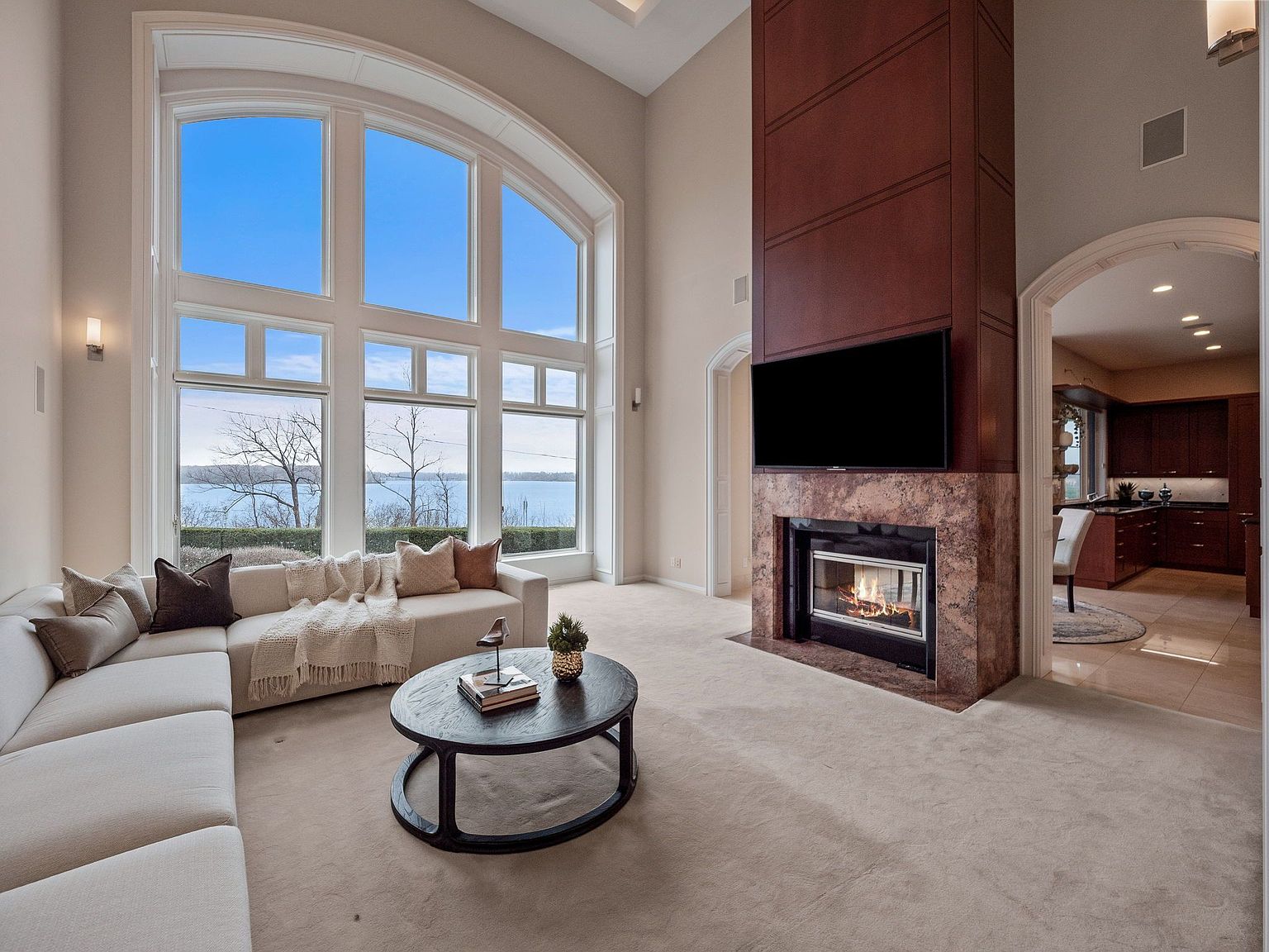 This is an interior shot of a luxurious living room featuring a large arched window with a scenic view of a lake. The room is furnished with a comfortable sectional sofa, a round coffee table, and a modern fireplace with a mounted television. The color palette is neutral, creating a bright and inviting atmosphere.
