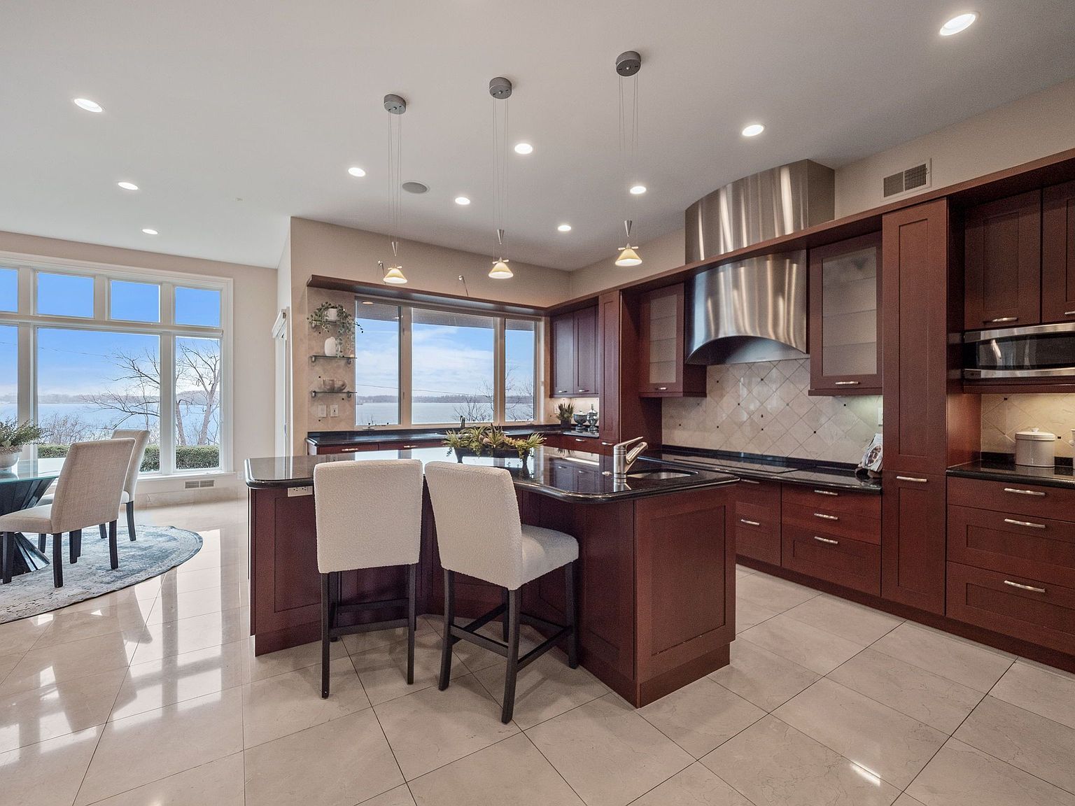 This is a spacious kitchen featuring dark wood cabinetry, black countertops, and stainless steel appliances. A large island with bar seating anchors the space, complemented by pendant lighting above. Large windows offer a scenic view, enhancing the kitchen's appeal.