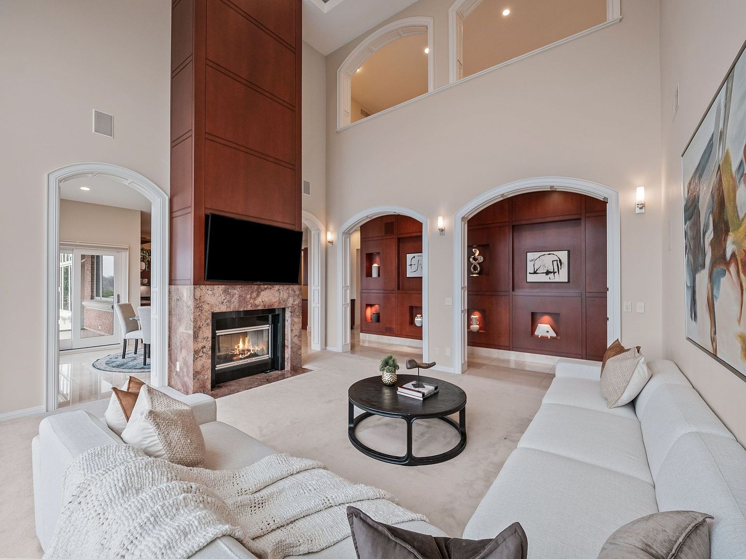 This is an interior shot of a luxurious living room featuring a tall fireplace with a mounted television, flanked by arched doorways. The room is furnished with a large, white sectional sofa and a round coffee table, all set on a neutral-toned carpet. The high ceilings and architectural details, such as the arched openings and wood paneling, contribute to an elegant and spacious atmosphere.