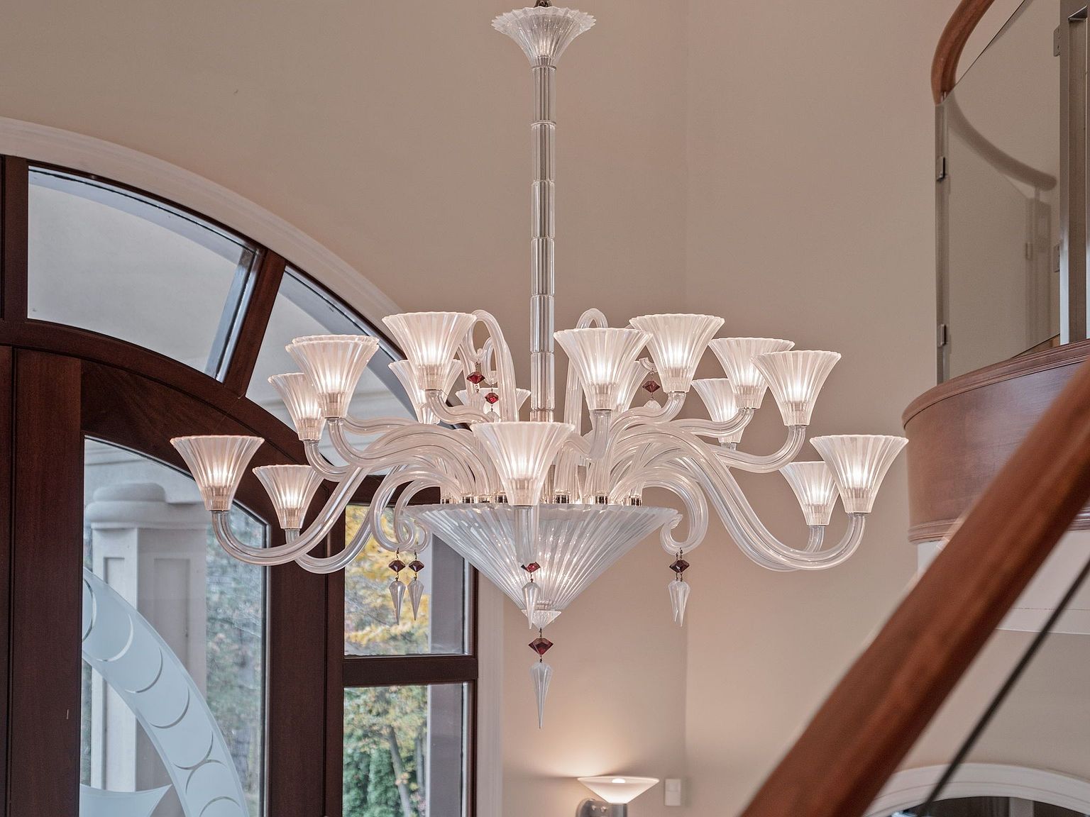 The image showcases an ornate, multi-tiered glass chandelier with delicate, fluted shades and decorative red accents. The fixture is suspended in a spacious interior, likely a foyer or grand hallway, with a glimpse of a curved staircase and a large arched window in the background. The chandelier's intricate design and the surrounding architectural details suggest a luxurious and elegant home.