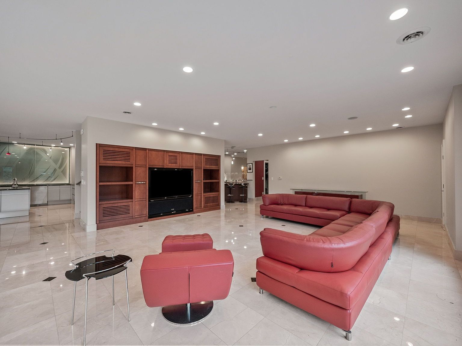 This is a spacious living room featuring a large red leather sectional sofa and a matching ottoman. A built-in wooden entertainment center houses a large television. The room has marble flooring and recessed lighting, creating a bright and open atmosphere.