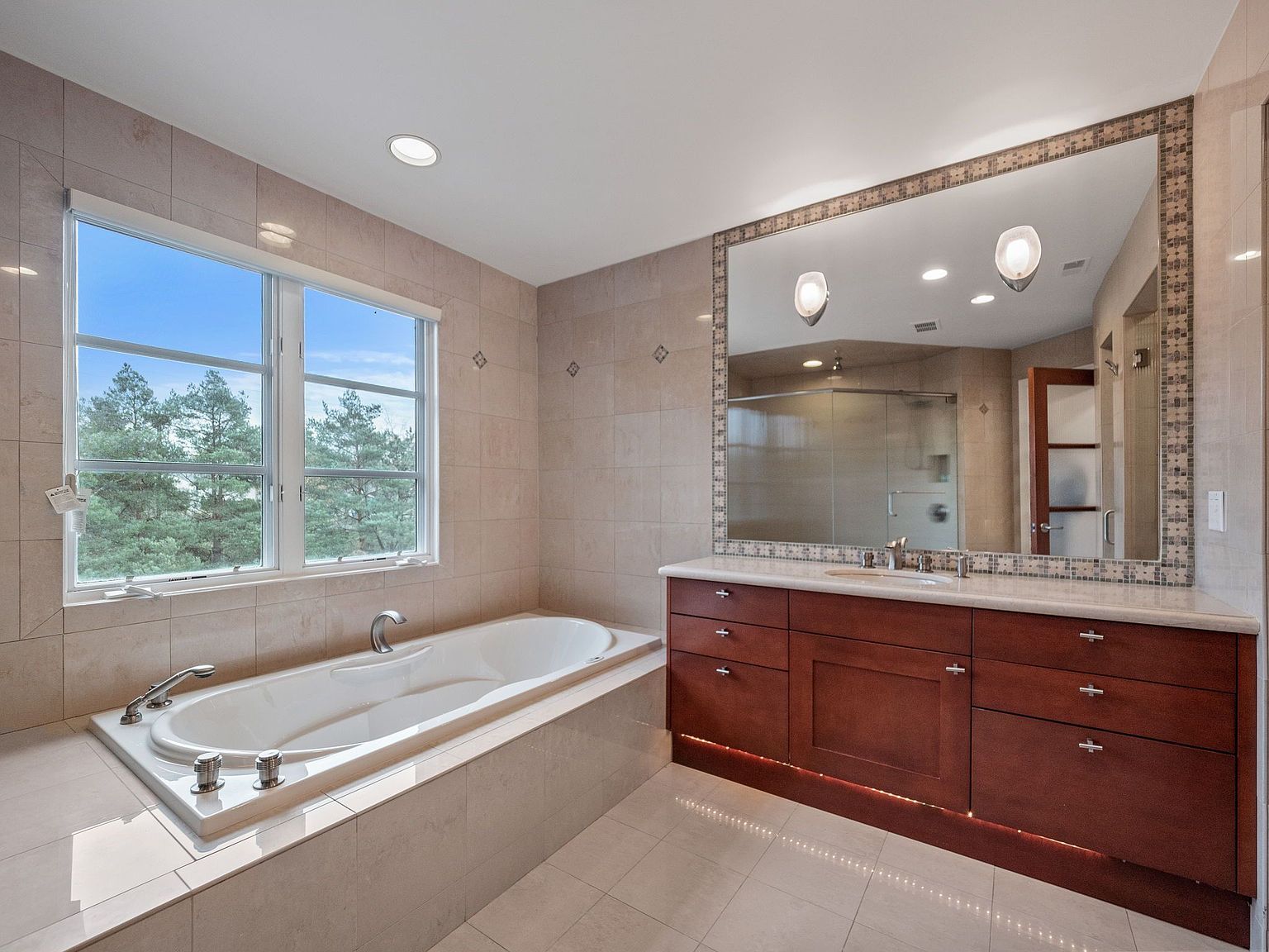 This is a well-lit primary bathroom featuring a large soaking tub set beneath a window offering a view of trees. A spacious vanity with dark wood cabinetry and a marble countertop is topped with a large mirror framed with mosaic tiles. The bathroom is tiled in neutral tones, creating a serene and luxurious atmosphere.