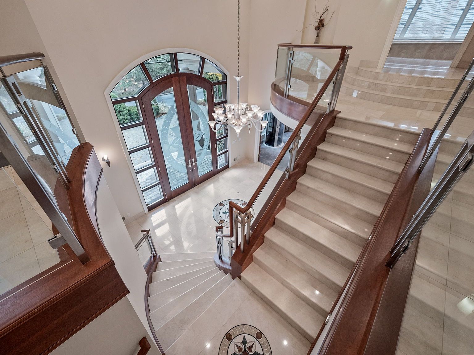 This is an interior shot of a grand foyer featuring a curved staircase with wooden handrails and glass panels. The entrance has large arched double doors with decorative glass, and a chandelier hangs from the ceiling. The flooring is light-colored tile with decorative inlays, creating an elegant and luxurious atmosphere.