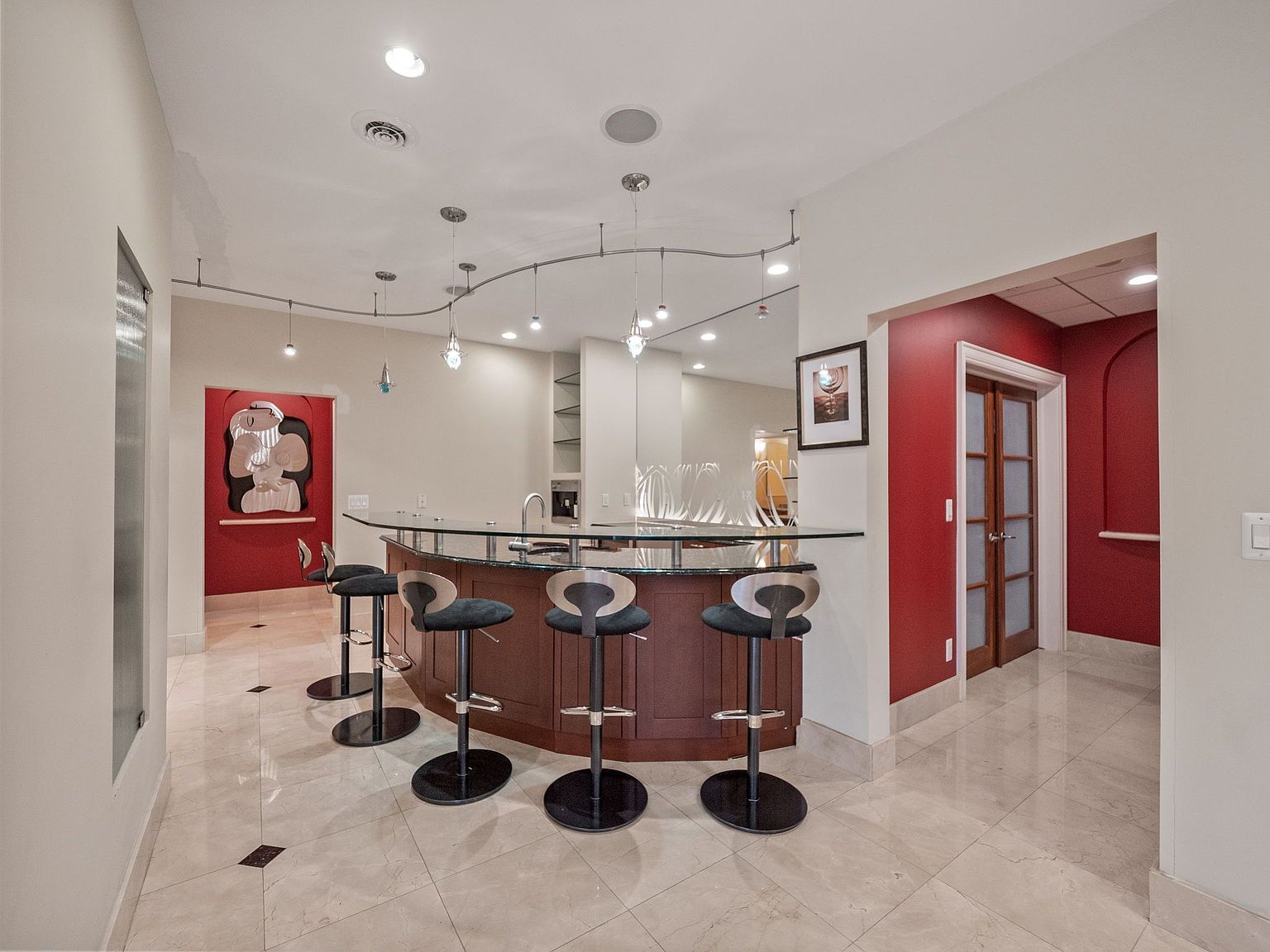 This interior shot showcases a stylish basement bar area, complete with a curved bar, glass countertop, and modern bar stools. The room features a red accent wall, recessed lighting, and a unique track lighting system above the bar, creating an inviting and sophisticated entertainment space. The marble tile flooring adds a touch of luxury.