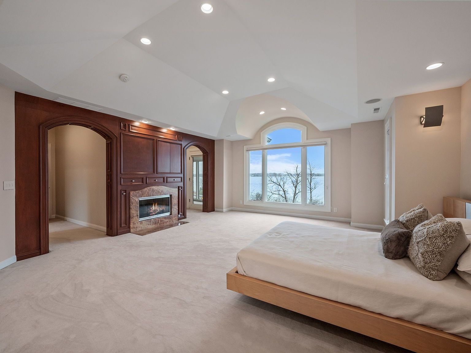 This is a spacious primary bedroom featuring a modern platform bed with neutral linens and decorative pillows. A prominent architectural feature is the dark wood built-in with a fireplace, flanked by arched doorways. Large windows offer a view of the water, and recessed lighting illuminates the room, creating a serene and luxurious atmosphere.