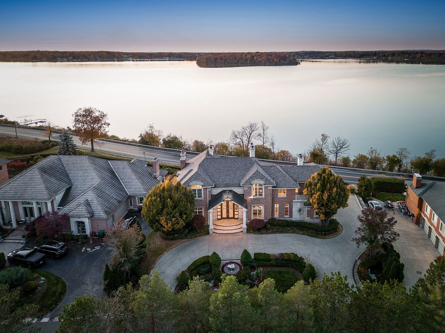This aerial view showcases a grand estate with meticulously landscaped grounds, a circular driveway, and multiple buildings, including a large main house and a separate garage or guest house. The property is situated on a waterfront, offering stunning lake views and a sense of privacy and luxury. The perspective is a high-angle shot, emphasizing the scale and beauty of the estate and its surroundings.