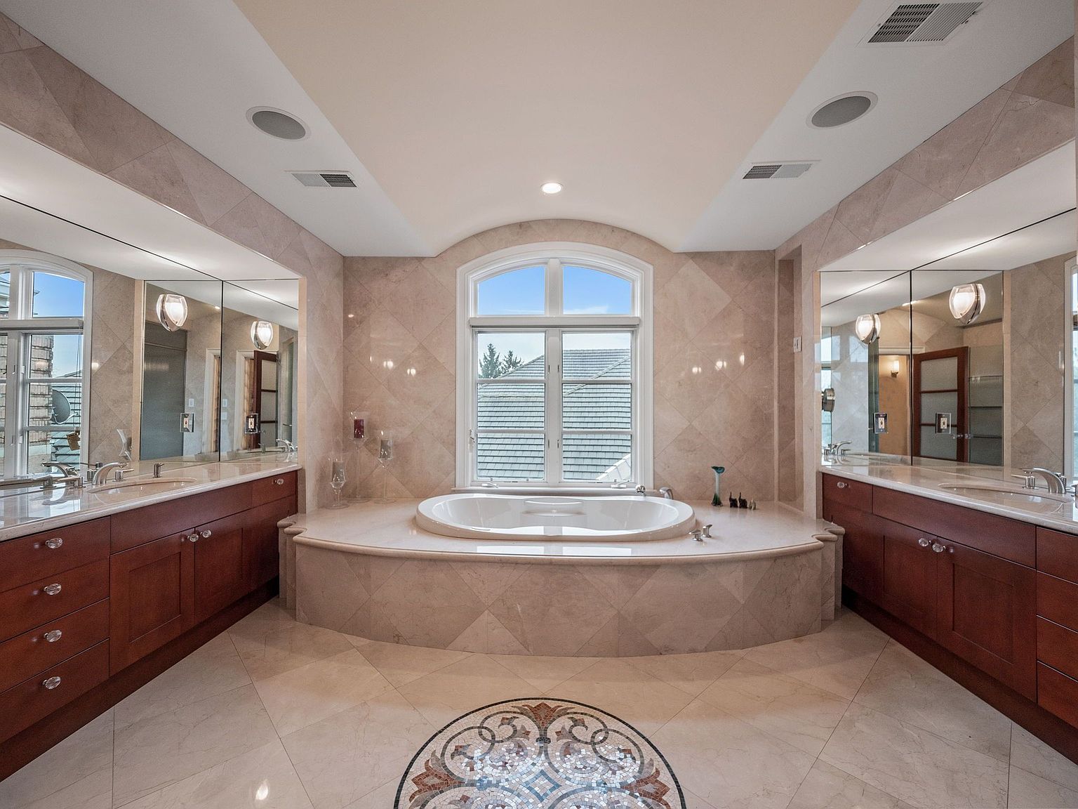 This is a luxurious primary bathroom featuring a large oval soaking tub centered beneath an arched window, providing natural light. The room is adorned with marble tiling on the walls and floor, complemented by dark wood vanities with dual sinks on either side. The overall impression is one of elegance and relaxation.