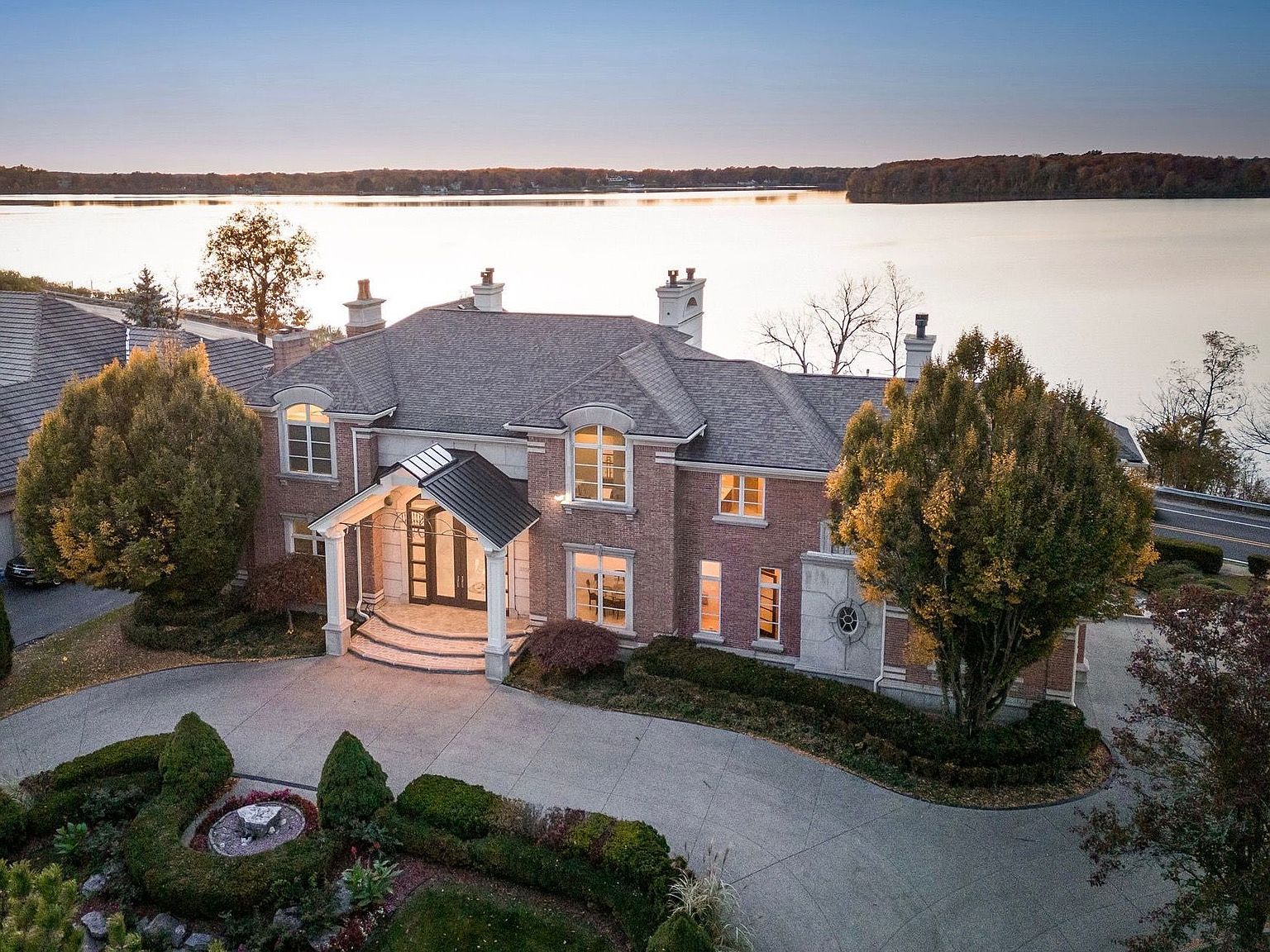 This is a stunning front view of a large, luxurious brick home situated on a waterfront property. The house features a grand entryway with a covered porch, elegant landscaping, and a circular driveway. The overall impression is one of opulence and sophisticated living, with the lake providing a picturesque backdrop.