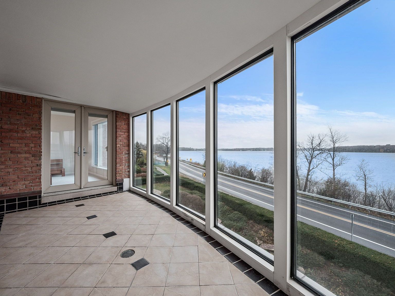 This is a sunroom or balcony featuring large windows that offer a panoramic view of a road, water, and sky. The flooring is tiled with dark accents, and the walls are a combination of brick and white framing. The space appears bright and airy, providing an excellent indoor-outdoor transition.