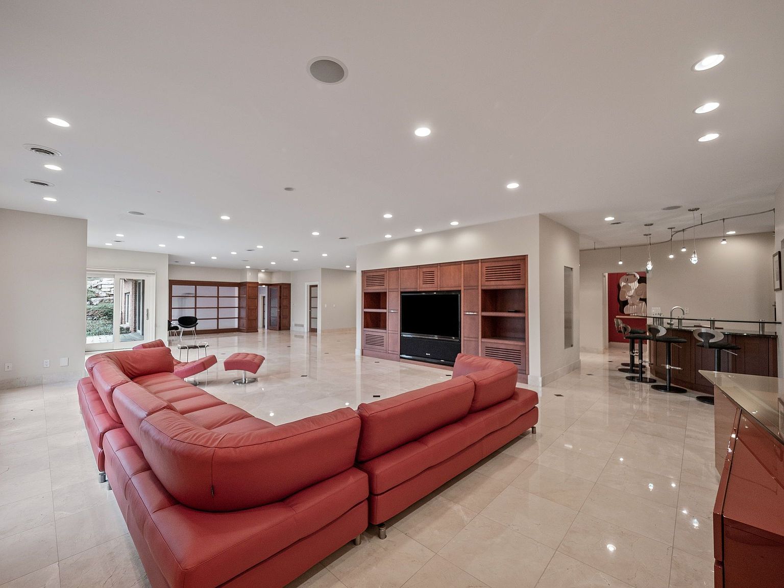 This is a spacious living room featuring a large, red, sectional sofa as the focal point. The room has a modern design with a built-in entertainment center, a bar area, and a neutral color palette. The flooring is a light-colored tile, and recessed lighting illuminates the space, creating a bright and inviting atmosphere.