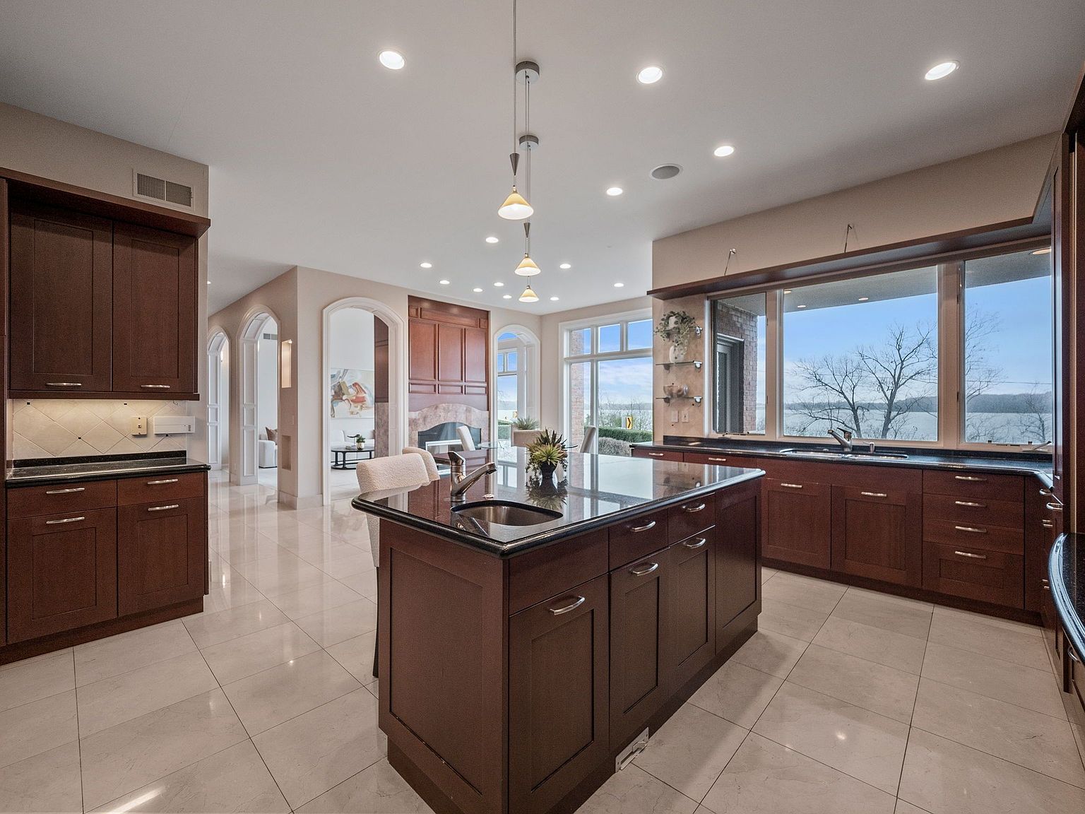 This is a spacious kitchen featuring dark wood cabinetry, black countertops, and a large island with a sink. The kitchen has a view of the water through a large window. The style is traditional and luxurious, with recessed lighting and a polished tile floor.