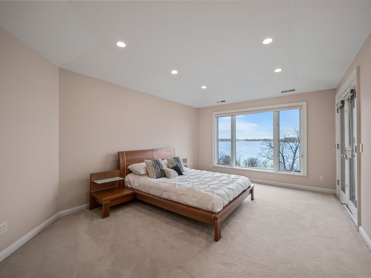 This is a bright and airy primary bedroom featuring a large window with a view of the water. The room is carpeted and has a neutral color palette, with a wooden bed frame and matching nightstand. Recessed lighting provides ample illumination, and a glass door leads to an outdoor space.