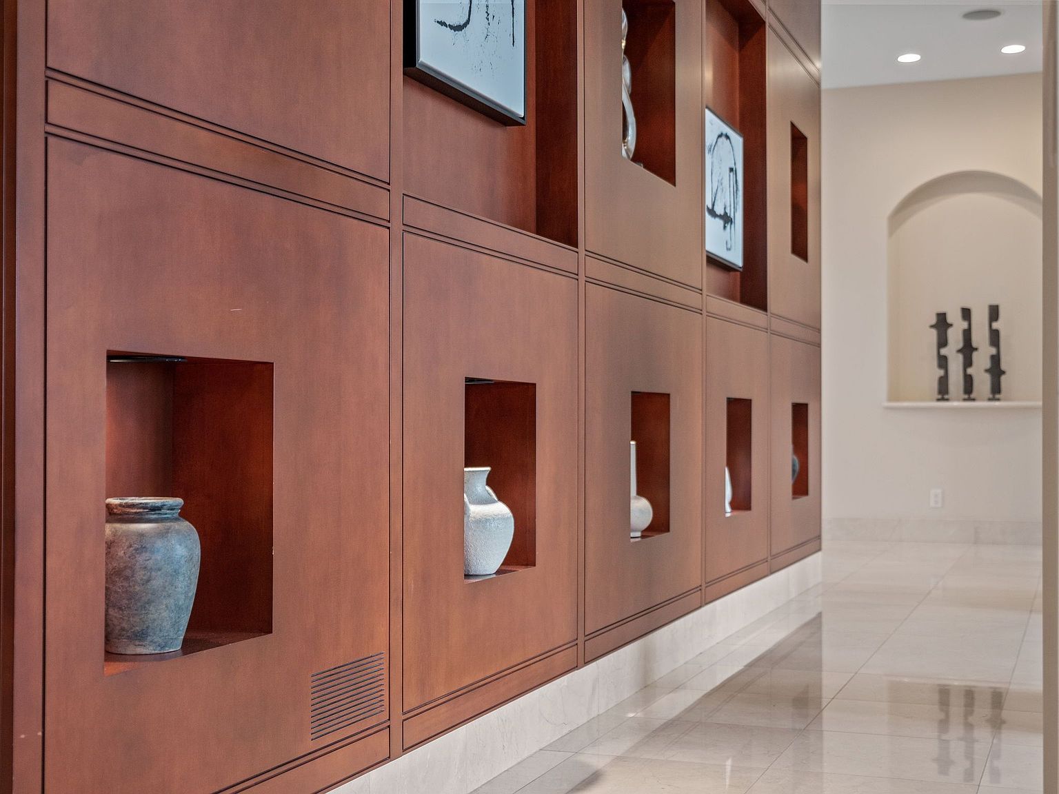 This interior shot showcases a hallway with a striking wall of built-in wooden shelving units, each displaying decorative vases and artwork. The hallway features light-colored tile flooring that reflects the ambient light, creating a bright and inviting space. The perspective emphasizes the depth of the hallway and the architectural details of the shelving.